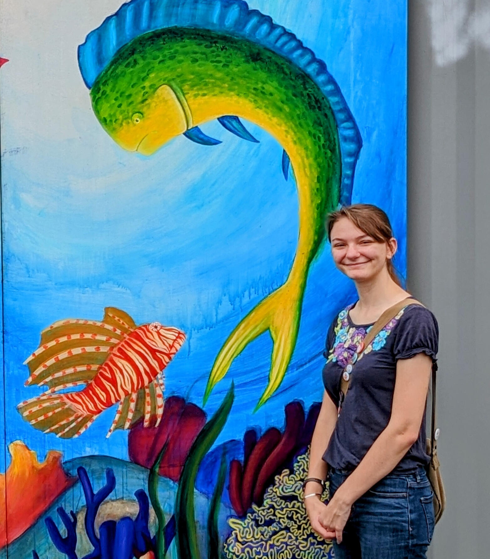 Greta Heitmueller standing in front of her 2022 high school senior final project mural. This mural is a 9 ft by 9 ft acrylic painting on wood attached to a shipping container depicting Florida sealife. There is a Mahi-mahi and a Lionfish.